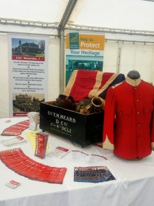 Cornwalls Regimental Museum on the Road! A display of items (including a flag, red military jacket and a bugle) arranged in a wooden chest, surrounded by leaflets, in a white marquee.