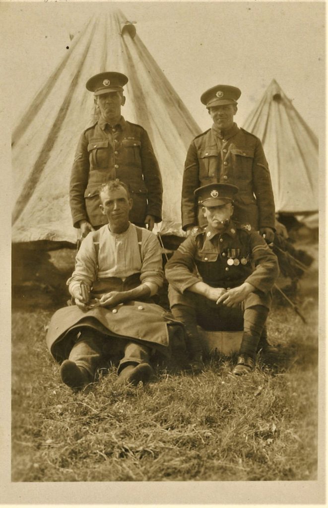 Photo of 4 non-commissioned officers – the man sitting on the left is using a sewing kit to fix the jacket which is lying across his lap