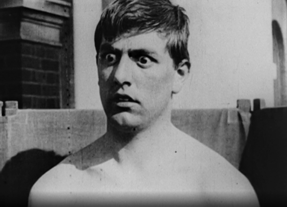 Black and white film still of a man’s head and shoulders. He looks to the left with wide eyes.