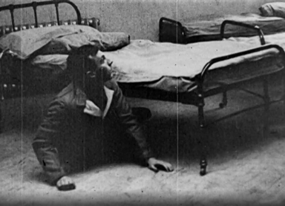 Black and white film still of a man peeking out from underneath a hospital bed.