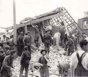 Black and white photograph of bomb damage in Bodmin with a crowd of people supporting a damaged structure.
