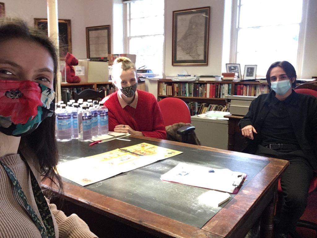 Two women and a man wearing covid-19 masks, distanced around a table. On top of the table lies a board game.