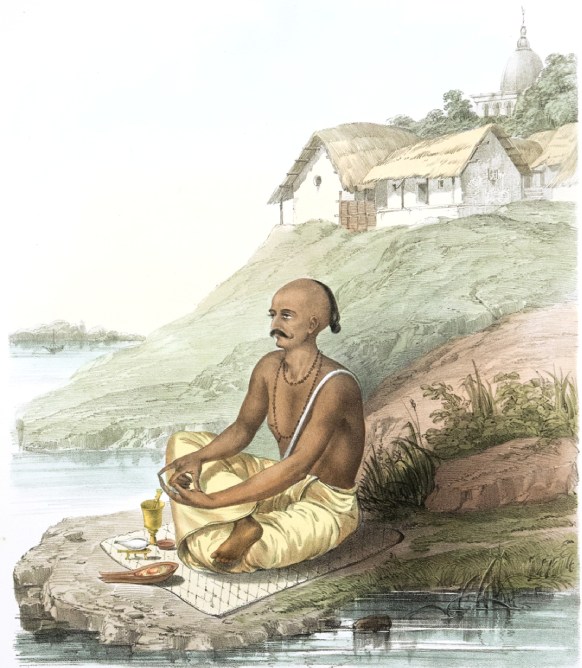 A man sat crossed legged on a mat by a river with a thatched roof house on a hill in the background.