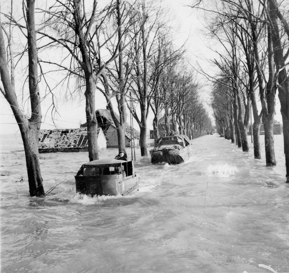 M29 Weasel followed by a DUKW amphibious vehicle navigate down a flooded avenue